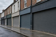 Synthetic editorial illustration. A row of brick and plaster shopfronts has closed metal shutters and blank fascias. The empty pavement and wet road recede under flat grey morning light.