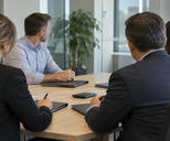 Synthetic editorial illustration. A small corporate finance meeting in an ordinary glass-walled office, anonymous treasury staff seen from behind with closed unmarked report folders and a face-down calculator, understated natural daylight.