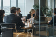 Synthetic editorial illustration. A documentary view through glass into a corporate board meeting with anonymous executives seated naturally around an ordinary table, papers seen from blank backs, no theatrical empty throne or handshake.