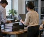 Synthetic editorial illustration. An ordinary registrar's office with anonymous colleagues sorting thick corporate record binders whose spines are blank and covers face the camera, subdued architectural context, no symbolic stock certificates.