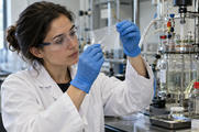 Synthetic editorial illustration. A materials researcher using gloved hands to inspect a small translucent polymer film at a practical laboratory bench, glass reactor and tubing in background, real tactile textures.