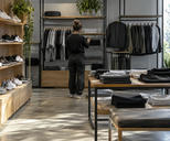 Synthetic editorial illustration. A quiet unbranded sportswear shop with orderly racks of plain trainers and athletic clothing and one shop worker seen from behind preparing the store, subdued natural daylight, no signage or discounts.
