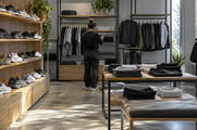 Synthetic editorial illustration. A quiet unbranded sportswear shop with orderly racks of plain trainers and athletic clothing and one shop worker seen from behind preparing the store, subdued natural daylight, no signage or discounts.