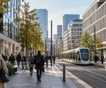 Synthetic editorial illustration. A wide documentary view of a generic continental-European business district and tram corridor during a normal morning commute, anonymous buildings and small pedestrians, no flags or recognizable monuments.