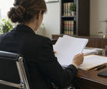Synthetic editorial illustration. A neutral ordinary institutional legal office with a lawyer seen from behind reviewing blank-backed case papers while an unbranded smartphone lies face-down nearby, everyday materials and soft window light.