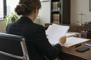 Synthetic editorial illustration. A neutral ordinary institutional legal office with a lawyer seen from behind reviewing blank-backed case papers while an unbranded smartphone lies face-down nearby, everyday materials and soft window light.