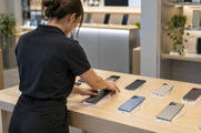 Synthetic editorial illustration. An anonymous electronics shop assistant viewed from behind carefully arranging generic unbranded smartphones face-down on an ordinary retail display, natural store lighting, no screens or price cards.