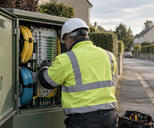 Synthetic editorial illustration. A telecom field technician seen from behind checking a fibre-optic distribution cabinet on a quiet suburban street, with realistic cable spools and tidy physical connections; no screen or markings visible.