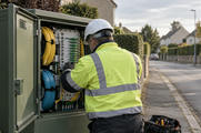 Synthetic editorial illustration. A telecom field technician seen from behind checking a fibre-optic distribution cabinet on a quiet suburban street, with realistic cable spools and tidy physical connections; no screen or markings visible.