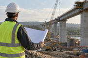 Synthetic editorial illustration. A civil engineer seen from behind reviewing an unmarked rolled plan at the edge of a working bridge construction site, credible concrete piers and construction equipment in the distance.