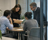 Synthetic editorial illustration. An anonymous financial-research team discussing an ordinary paper folder in a small glass meeting area, medium-wide observational composition, practical office texture and no visible chart or text.