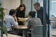 Synthetic editorial illustration. An anonymous financial-research team discussing an ordinary paper folder in a small glass meeting area, medium-wide observational composition, practical office texture and no visible chart or text.