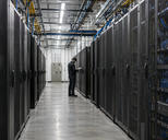 Synthetic editorial illustration. A working high-density computing facility with realistic server racks and practical overhead cabling, a small unidentifiable technician inspecting the aisle, no glowing sci-fi chips or visible screens.