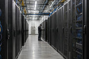 Synthetic editorial illustration. A working high-density computing facility with realistic server racks and practical overhead cabling, a small unidentifiable technician inspecting the aisle, no glowing sci-fi chips or visible screens.