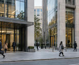 Synthetic editorial illustration. A street-level documentary view of two adjacent anonymous financial-office buildings with a few commuters, neutral stone and glass façades, no logos, bank names or symbolic miniature buildings.