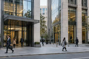 Synthetic editorial illustration. A street-level documentary view of two adjacent anonymous financial-office buildings with a few commuters, neutral stone and glass façades, no logos, bank names or symbolic miniature buildings.