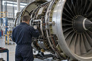 Synthetic editorial illustration. A technician seen from behind inspecting a credible aircraft-engine assembly in a functioning aerospace workshop, real machined metal components and practical fixtures, natural industrial daylight.
