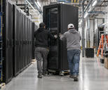 Synthetic editorial illustration. A new data-centre equipment hall during normal installation, two small technicians seen from behind guiding a real rack cabinet into place, practical cables and believable industrial scale.