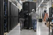 Synthetic editorial illustration. A new data-centre equipment hall during normal installation, two small technicians seen from behind guiding a real rack cabinet into place, practical cables and believable industrial scale.