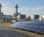 Synthetic editorial illustration. A credible electricity-generating site with ordinary gas-turbine buildings and a modest field of solar panels adjacent, documentary scale and distant worker, balanced natural daylight, no surreal juxtaposition.