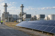 Synthetic editorial illustration. A credible electricity-generating site with ordinary gas-turbine buildings and a modest field of solar panels adjacent, documentary scale and distant worker, balanced natural daylight, no surreal juxtaposition.