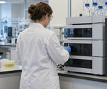 Synthetic editorial illustration. A pharmaceutical researcher seen from behind working at a credible analytical laboratory instrument with closed sample compartments, calm workaday atmosphere, no display or paperwork text.