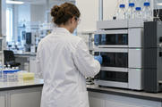 Synthetic editorial illustration. A pharmaceutical researcher seen from behind working at a credible analytical laboratory instrument with closed sample compartments, calm workaday atmosphere, no display or paperwork text.