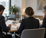 Synthetic editorial illustration. An anonymous investment committee working around a modest table in a contemporary office, three colleagues seen obliquely from behind reviewing blank-backed papers, restrained observational lighting.