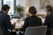 Synthetic editorial illustration. An anonymous investment committee working around a modest table in a contemporary office, three colleagues seen obliquely from behind reviewing blank-backed papers, restrained observational lighting.