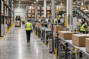 Synthetic editorial illustration. A documentary view across a functioning anonymous distribution warehouse with orderly machinery and a small manager walking the aisle, representing the operating businesses behind private-market portfolios.