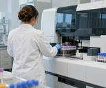 Synthetic editorial illustration. A clinical diagnostics laboratory with an anonymous technician seen from behind tending a credible automated sample analyser, orderly racks of sealed tubes, no visible labels or computer screens.