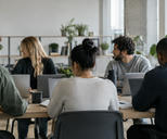 Synthetic editorial illustration. An anonymous software team collaborating in a plain modern workspace with laptop backs toward camera and all screens hidden, relaxed natural working postures, credible office depth and daylight.