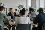 Synthetic editorial illustration. An anonymous software team collaborating in a plain modern workspace with laptop backs toward camera and all screens hidden, relaxed natural working postures, credible office depth and daylight.