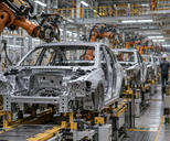 Synthetic editorial illustration. A modern unbranded automotive plant with unfinished vehicle bodies moving along a credible assembly line, practical robotic arms and distant workers seen from behind, no product logos.