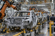 Synthetic editorial illustration. A modern unbranded automotive plant with unfinished vehicle bodies moving along a credible assembly line, practical robotic arms and distant workers seen from behind, no product logos.
