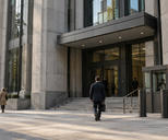 Synthetic editorial illustration. An anonymous public legal-administration building exterior at street level with small pedestrians, sober stone-and-glass architecture and a subtle unmarked security entrance, no gavel props or court symbols.
