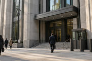 Synthetic editorial illustration. An anonymous public legal-administration building exterior at street level with small pedestrians, sober stone-and-glass architecture and a subtle unmarked security entrance, no gavel props or court symbols.