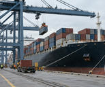 Synthetic editorial illustration. Working container port from quayside, a generic container ship and gantry cranes unloading plain containers, distant small dockworkers, soft overcast daylight and believable industrial scale.