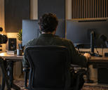 Synthetic editorial illustration. A real unbranded audio-and-video production editing workspace with a small camera, microphone and sound panels, a producer seen from behind and all monitor screens turned away.