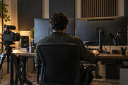 Synthetic editorial illustration. A real unbranded audio-and-video production editing workspace with a small camera, microphone and sound panels, a producer seen from behind and all monitor screens turned away.