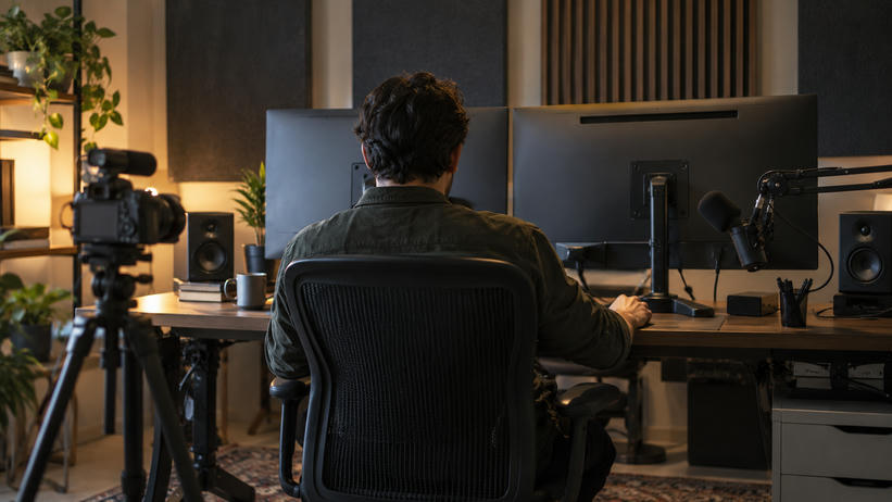 Synthetic editorial illustration. A real unbranded audio-and-video production editing workspace with a small camera, microphone and sound panels, a producer seen from behind and all monitor screens turned away.