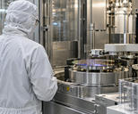Synthetic editorial illustration. A semiconductor-equipment engineer seen from behind beside a credible precision wafer-handling machine in a clean fabrication room, stainless enclosures and practical robotics, no screens.