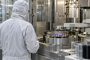 Synthetic editorial illustration. A semiconductor-equipment engineer seen from behind beside a credible precision wafer-handling machine in a clean fabrication room, stainless enclosures and practical robotics, no screens.