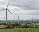 Synthetic editorial illustration. A natural documentary landscape showing a working onshore wind farm and electricity transmission connections amid agricultural fields, plausible turbine scale and soft overcast light, no symbolic eco props.