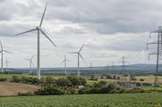 Synthetic editorial illustration. A natural documentary landscape showing a working onshore wind farm and electricity transmission connections amid agricultural fields, plausible turbine scale and soft overcast light, no symbolic eco props.