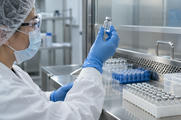 Synthetic editorial illustration. An anonymous pharmaceutical quality laboratory with a gloved scientist inspecting a sealed clear vial at a stainless bench, neatly ordered sample racks and clean equipment, no labels or syringes.