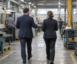 Synthetic editorial illustration. An anonymous industrial facility with two business representatives seen from behind touring a practical manufacturing floor, real machinery and worn working surfaces, documentary medium-wide framing.