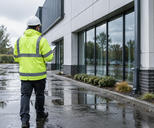 Synthetic editorial illustration. An anonymous insurance surveyor seen from behind inspecting a rain-soaked commercial building exterior after normal heavy rain, reflective puddles and credible architecture, no disaster dramatisation.