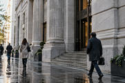 Synthetic editorial illustration. A sober anonymous institutional financial building at street level after rain, a few small pedestrians and real stone textures, balanced morning light, no flags, names or recognizable landmark.