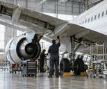 Synthetic editorial illustration. An anonymous aircraft maintenance hangar with a realistic unmarked narrow-body passenger aircraft and small mechanics seen from behind beneath the wing, industrial scale and diffuse daylight.